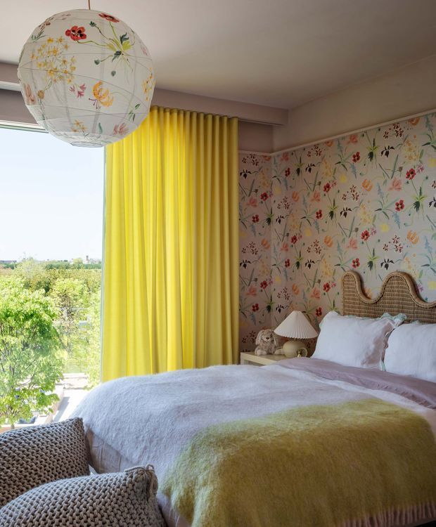 A bright, playful bedroom with a large bed dressed in white and pale yellow bedding, a woven rattan headboard, floral botanical wallpaper, and vivid yellow floor-to-ceiling curtains beside a large window. A floral paper pendant light and textured knitted seating add layered decorative detail. The room overlooks leafy greenery and an open landscape.