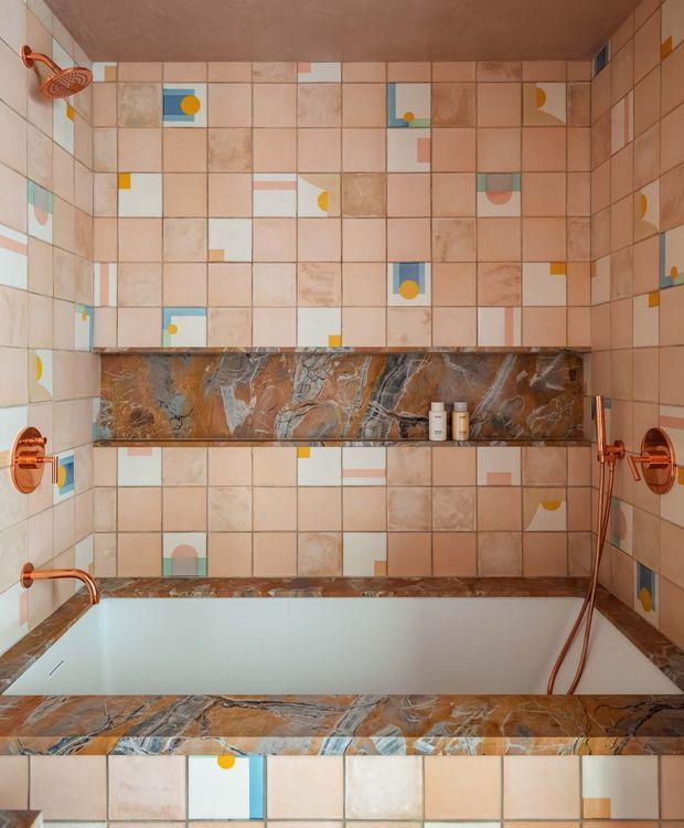 A close-up view of a highly tiled bathroom featuring a built-in bathtub, wall-mounted shower and copper-toned fixtures. The walls are covered in pale blush and beige square tiles with scattered geometric and abstract accent tiles. A dramatic brown, grey and blue veined stone shelf and bath surround runs horizontally through the space, complemented by matching stone detailing near the floor. The overall composition is graphic, colourful and retro-inspired, with a warm palette and reflective surfaces.