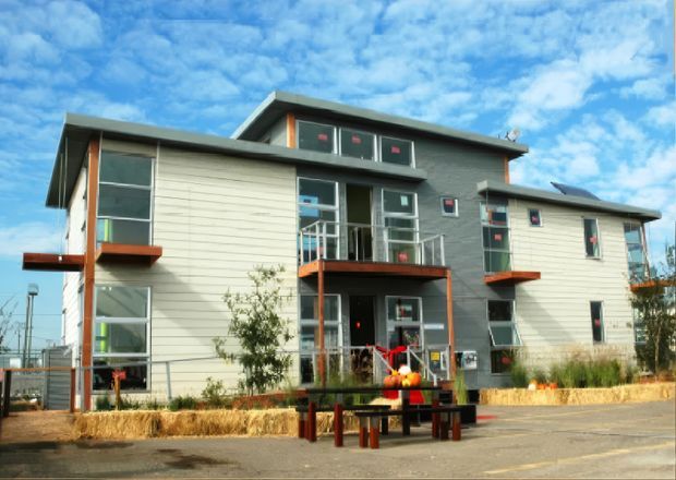 Exterior view of a contemporary multi-storey residential building with asymmetrical rooflines, extensive glazing, light horizontal cladding, dark grey upper-level cladding, and red-orange balcony and trim accents. The building sits beside a paved communal area with landscaping and outdoor seating.