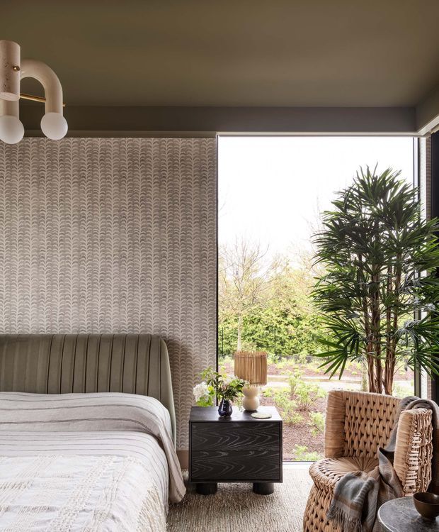 A softly lit bedroom with a large floor-to-ceiling window overlooking a planted garden. The room features a curved upholstered bedhead, patterned neutral wallpaper, layered white and grey bedding, a dark wood bedside table, a woven rattan armchair, indoor tropical plants, and sculptural pendant lighting. The atmosphere is warm, refined, and calm, with a contemporary organic feel.
