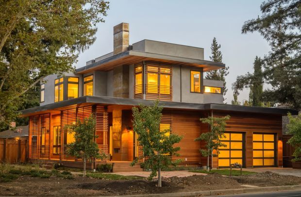Two-storey contemporary residential house with a flat-roofed, box-like composition, warm timber cladding, smooth grey rendered walls, extensive horizontal timber sun-shading, and glowing amber windows. A prominent chimney rises above the upper level, while an attached single-car garage occupies the right side. Mature trees and landscaped planting frame the facade.