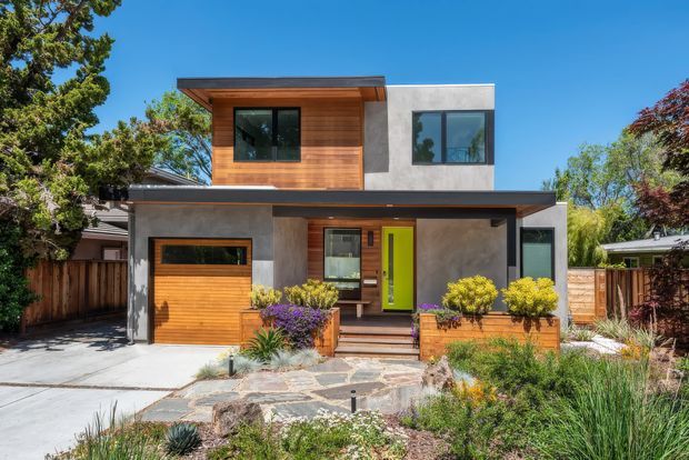 Front exterior view of a contemporary two-storey house with a strong boxy form, flat rooflines, grey rendered walls, warm horizontal timber cladding, large dark-framed windows, and a bright lime-green entry door. A timber garage door, paved driveway, layered native-style landscaping, timber fencing, and stone stepping paths are visible.
