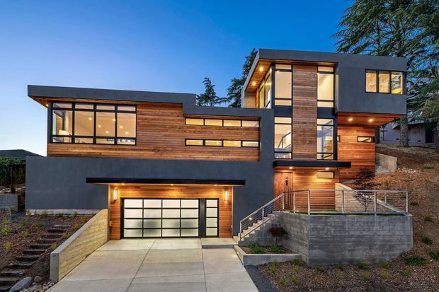 Photograph of a modern multi-level residence at dusk, featuring a strong geometric composition, flat rooflines, dark charcoal rendered walls, warm timber cladding, extensive black-framed glazing, a double garage, and an elevated entry terrace accessed by stairs.