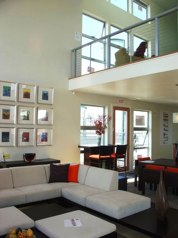 A bright, double-height contemporary lounge and reception-like interior with a large white sectional sofa, black coffee table, red and black accent cushions, framed artwork, and a dark dining or meeting table with red chairs. Tall windows and an upper-level mezzanine with glass balustrading bring in abundant natural light. A small high table and chairs sit near the glazed entrance, while decorative vases and artwork add visual interest.