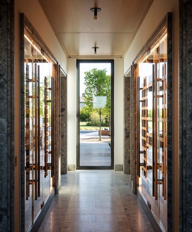 A symmetrical, narrow interior entry hall framed by two large glazed built-in display or storage walls with dark stone surrounds and warm timber detailing. The hall has a polished stone-tile floor, a light panelled ceiling with minimalist pendant fixtures, and a full-height glazed door at the far end opening toward an outdoor walkway and landscaped street setting. The composition is refined and architectural, with strong axial perspective and abundant natural light.