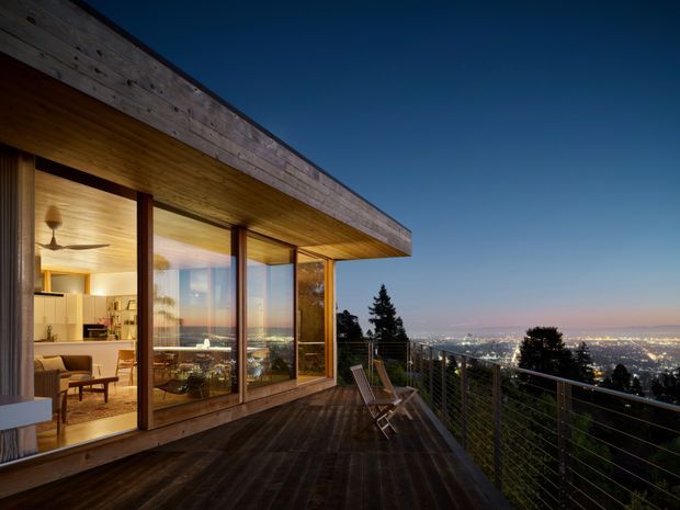 Twilight exterior view of a modern timber-clad home with a broad projecting flat roof, extensive floor-to-ceiling glazing, and a large dark timber deck with cable railing. The open-plan living and dining area is visible through the glazing, with warm timber ceilings and built-in joinery. The home is elevated on a sloping site overlooking a brightly lit urban landscape.