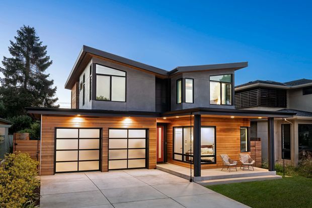 Exterior architectural photograph of a contemporary two-storey home with strong horizontal roof planes, dark grey upper-level cladding, warm timber garage and entry cladding, large black-framed windows, two glazed garage doors, and a covered ground-floor terrace. The house is set in a suburban residential context with a concrete driveway and lawn.
