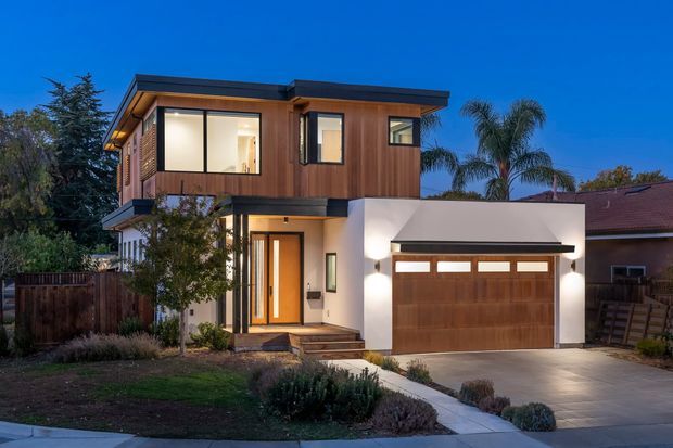 Twilight exterior view of a contemporary two-storey suburban home. The house has a projecting upper level clad in warm vertical timber, a flat roof, large upper windows, a white rendered lower facade, a timber front door, and an integrated double garage with a wood-look door. A landscaped front garden and driveway frame the facade.
