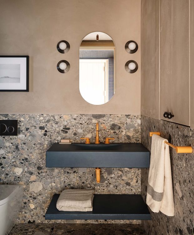 Compact, richly finished bathroom with a wall-mounted matte black basin and matching floating bench, surrounded by dramatic terrazzo-style stone wall and floor surfaces. Warm copper-toned tapware, exposed drain, towel rail, and flush plate provide accents against the dark and mottled stone. An arched pill-shaped mirror, paired round wall lights, and framed artwork complete the refined contemporary scheme.