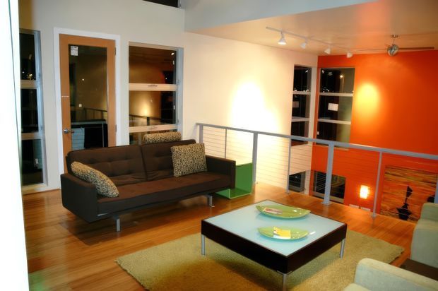 A bright contemporary living room in a split-level or mezzanine-style home, with warm timber flooring, a dark brown sofa, patterned cushions, a low square coffee table, and a pale rug. A grey metal balustrade overlooks a lower level, while vivid orange feature walls, built-in shelving, recessed lighting, and large internal openings create a bold modern interior.