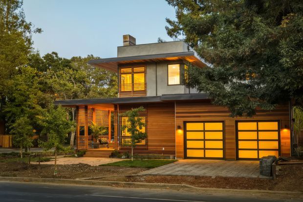 A two-storey contemporary residential house viewed from the street, set among mature trees. The home has a flat roof, projecting upper-level form, warm horizontal timber wall cladding, dark-framed windows with timber louvres, and two illuminated sectional garage doors. A paved driveway and small landscaped front garden sit along the road.