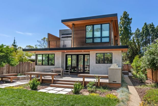 Modern two-storey residence viewed from the landscaped backyard, featuring warm timber upper-level cladding, smooth grey rendered walls, expansive dark-framed glazing, and broad flat roof overhangs. The ground level opens onto a paved outdoor terrace with built-in bench seating, a dining table, lounge chairs, and a built-in outdoor grill. Timber fencing, planted garden beds, lawn, and mature trees surround the contemporary home.