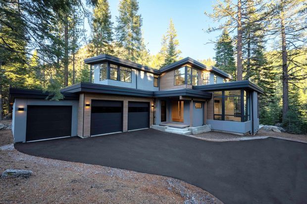 Exterior view of a contemporary multi-level house set among tall forest trees. The home has dark horizontal cladding, extensive black-framed glazing, several projecting roof volumes, a covered entry, and a broad dark paved driveway leading to three garage doors.