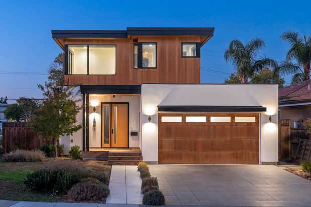 Street-facing view of a contemporary two-storey residence at dusk, featuring a warm timber-clad upper level, white-rendered lower facade, a projecting low-pitched roof, large upper-level windows, a timber front door, and an integrated double garage with a wood-panel garage door. The front garden and paved driveway are visible.