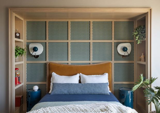 A carefully styled bedroom with a built-in timber-framed wall spanning the bed. The wall features dark patterned inset panels, integrated open shelving with books and decorative objects, two wall-mounted spherical lights, and indoor plants. A mustard-brown upholstered headboard contrasts with blue bedding, white pillows, and a textured cream throw. The room has a warm, layered, eclectic yet refined character.