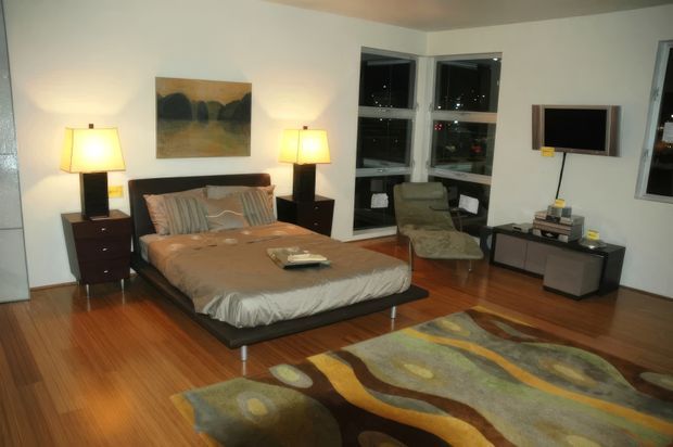 A modern bedroom with a low platform bed, dark wood bedside tables, matching table lamps, a wall-mounted television, a compact lounge chair, and a long low media console. Warm timber flooring and a large abstract-patterned rug add colour and texture, while corner windows provide nighttime city views.