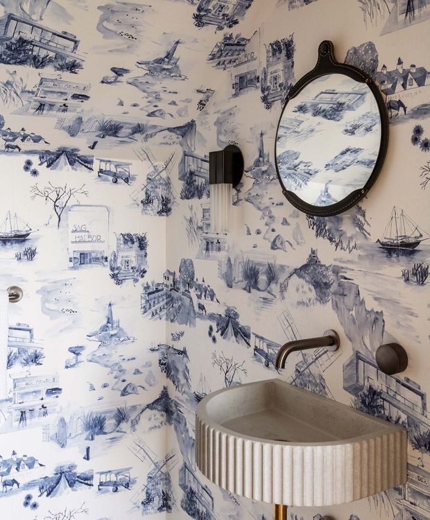 A compact bathroom with a sculptural fluted wall-mounted basin, dark metal wall-mounted tapware, and an oval framed mirror. The walls are covered in expressive blue-and-white illustrated scenic wallpaper depicting coastal and village scenes. The space has a playful, eclectic, highly decorative character with a warm neutral basin and dark contrasting fixtures.