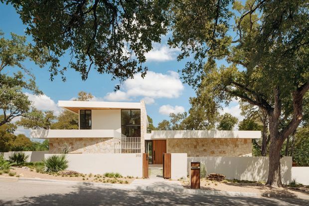 Street-facing view of a low-slung contemporary residence set among mature trees. The house features a flat roof, strong horizontal lines, pale rendered walls, natural light-coloured stone cladding, expansive dark-framed glazing, a timber entry gate, and a minimalist landscaped frontage.