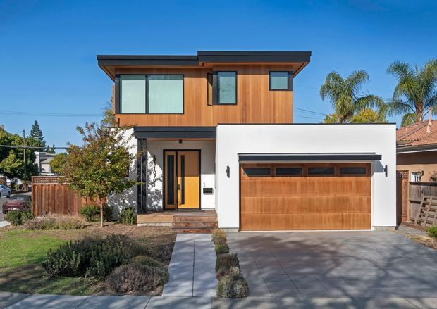 Front exterior view of a contemporary two-storey urban home. The facade combines warm vertical timber cladding on the upper level and garage door with crisp white rendered walls, dark metal roof and trim, large windows, and an attached single-car garage. A concrete driveway and walkway lead through a modest landscaped front garden.