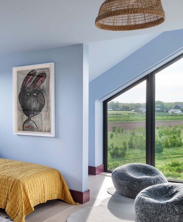 A bright bedroom with pale blue walls, a mustard quilted bedspread, and two rounded textured lounge chairs positioned beside expansive black-framed angled glazing. A woven rattan pendant hangs from the ceiling, while a framed abstract artwork decorates the wall. The windows overlook a green rural landscape, and deep burgundy skirting accents the room.