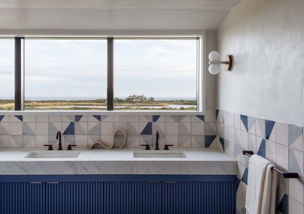 A bright contemporary bathroom with a long double-sink vanity beneath a wide picture window overlooking a coastal landscape. The room features a white marble-look benchtop, deep navy fluted cabinetry, geometric patterned wall tiles in white, grey, navy and muted terracotta, dark tapware, and a sculptural wall light. A towel rail with folded towels is visible at right.