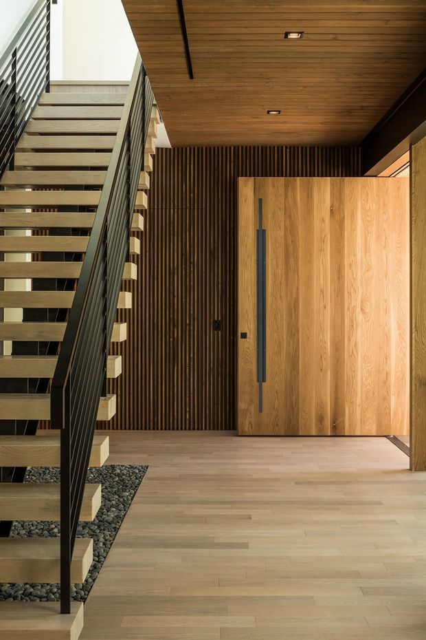 Contemporary residential entry hall featuring an open-riser timber staircase with dark metal balustrades, light timber flooring, a large timber pivot entry door, and a dramatic vertical timber batten feature wall. Dark pebble-filled recesses beneath the stairs add texture, while recessed ceiling lighting and warm wood tones create a refined, natural atmosphere.