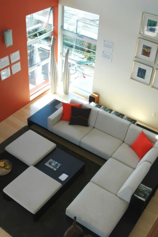 A bright, contemporary living room viewed from above, featuring a large light-grey L-shaped sectional sofa with red and black accent cushions, a black rectangular coffee table, and matching ottomans on a dark rug. Tall corner windows with sheer curtains bring in abundant daylight, while one wall is painted a bold terracotta-red and the adjacent wall displays framed artwork and notices. Light timber flooring and clean-lined furnishings create a modern, graphic interior.