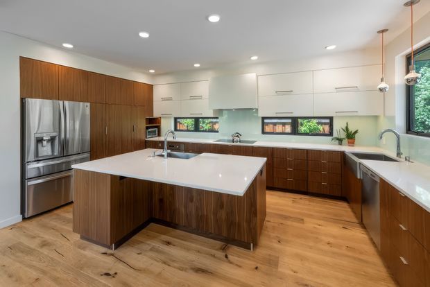 Bright, spacious contemporary kitchen with a large central island, extensive white and dark timber cabinetry, white stone benchtops, stainless-steel appliances, timber flooring, integrated lighting, and multiple windows providing natural light.