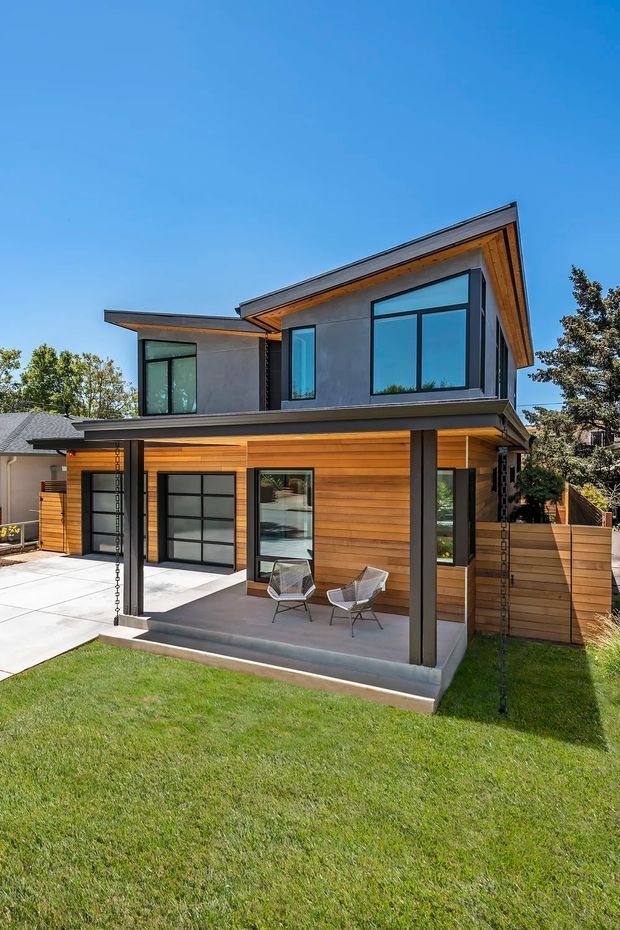 Exterior view of a contemporary two-storey residential extension or compact home, featuring warm horizontal timber cladding on the ground floor, grey rendered upper walls, expansive black-framed glazing, a shallow asymmetrical pitched roof with exposed timber eaves, and a covered patio with two outdoor chairs. A garage and paved driveway are visible alongside the landscaped lawn.