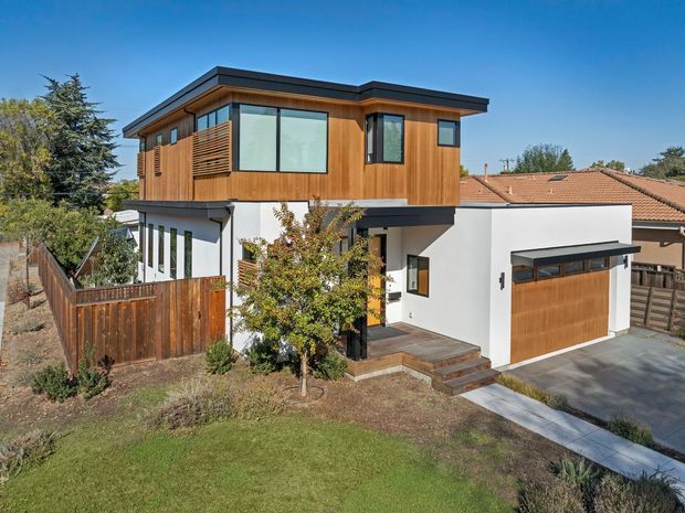 Exterior view of a contemporary two-storey residence with a prominent upper-level timber-clad volume, crisp white rendered lower walls, broad black roof edging, large glazed windows, and an integrated garage. The house sits on a landscaped suburban site with a timber boundary fence, front lawn, paved entry path, and neighbouring homes visible behind it.