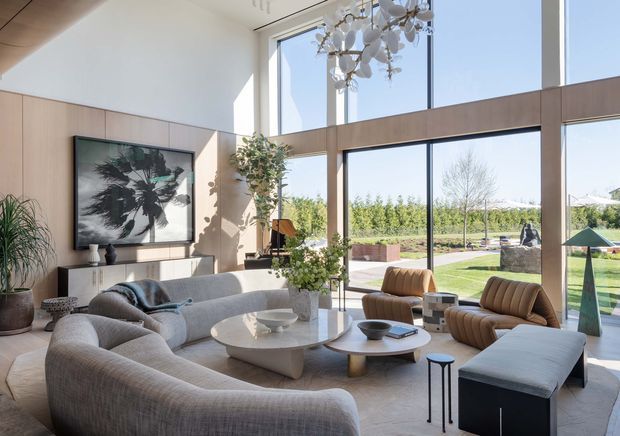 Bright, spacious contemporary living room with double-height floor-to-ceiling glazing overlooking a landscaped garden. The room features curved upholstered sofas, sculptural coffee tables, leather lounge chairs, a large abstract artwork, indoor plants, and a statement chandelier.