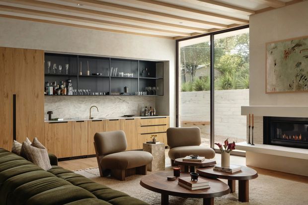 Warm contemporary open-plan living room with an integrated bar and kitchenette along the rear wall, featuring light timber cabinetry, dark open shelving, a pale stone or marble-look backsplash, and a compact brass-toned sink tap. The room has a large olive-green sectional sofa, sculptural brown lounge chairs, layered round timber coffee tables, and a linear fireplace set into a pale built-in wall. Full-height black-framed glazing opens visually to a planted courtyard. Exposed light timber ceiling beams and a neutral textured palette create a refined, relaxed atmosphere.