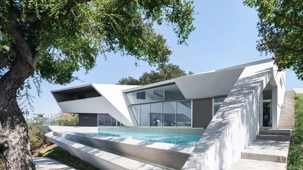 A striking modern white geometric house set among mature trees, viewed from the pool terrace. The building features sharply angled cantilevered forms, extensive floor-to-ceiling glazing, dark recessed openings, and a long narrow swimming pool with sculptural stepped edges. Exterior stairs lead up along the right side, while landscaped surroundings and neighboring structures are visible in the background.