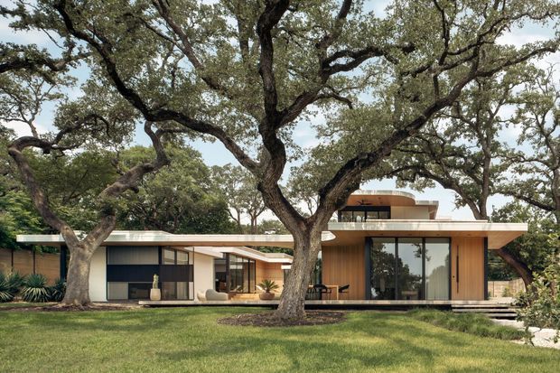A low-slung modern single-storey residence set among mature spreading oak trees. The house features broad horizontal rooflines, warm timber-clad walls, extensive dark-framed glazing, crisp pale exterior surfaces, and a layered indoor-outdoor relationship opening onto a lawn and terrace.