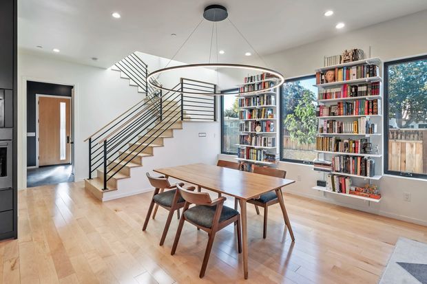 Bright open-plan dining and reading area with a rectangular timber dining table and mid-century style chairs, pale timber flooring, extensive built-in and freestanding bookshelves, and a sculptural circular pendant light. A contemporary open-riser staircase with black metal railings leads upward beside large windows, with a hallway and timber door visible beyond.