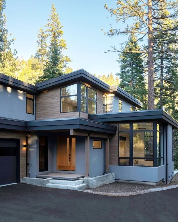 Exterior view of a contemporary multi-level house set among tall pine trees. The building has a low-pitched, layered roofline, dark metal fascia, grey rendered walls, warm horizontal timber cladding, large black-framed windows, and a recessed timber entry door. A paved driveway and gravel edging are visible in the foreground.