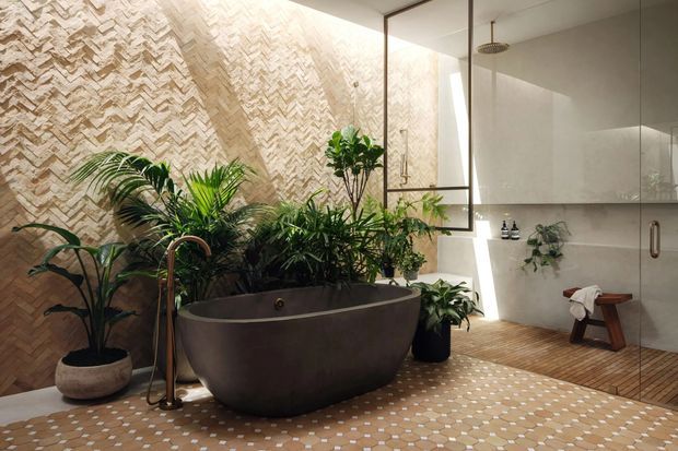 A warm, spa-like bathroom featuring a freestanding dark stone or composite bathtub surrounded by lush tropical plants. The room has a textured chevron-patterned stone feature wall, a glass-enclosed walk-in shower with a rainfall showerhead, wood-look slatted flooring in the shower area, and small geometric patterned floor tiles throughout. Natural light enters through a narrow vertical opening, creating a calm biophilic atmosphere.