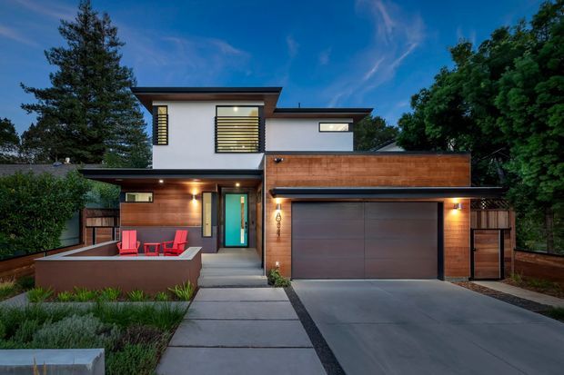 Contemporary two-storey residential facade at dusk, featuring a flat-roofed composition, white rendered upper walls, warm timber-toned cladding, dark-framed windows with horizontal screening, an integrated double garage, and a concrete driveway and entry path. Landscaping and a small front terrace soften the street-facing elevation.
