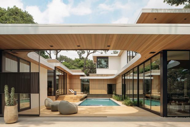 Contemporary courtyard residence organized around a compact swimming pool, with low flat roof planes, extensive floor-to-ceiling glazing, warm timber soffits and decking, pale rendered walls, and strong black metal framing. Sculptural outdoor seating and planters sit beside the pool, while mature trees are visible beyond the enclosed courtyard.