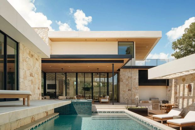 A bright contemporary courtyard-style residence with strong indoor–outdoor connections. The composition centres on a swimming pool adjoining a glazed living area, with pale irregular stone walls, warm timber soffits, broad flat roof planes, black-framed glazing, and a glass balustrade. White built forms and timber-lined overhangs create a clean, resort-like architectural setting with outdoor lounge furniture around the pool.