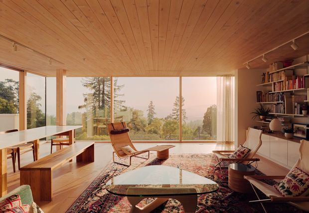 Warm mid-century-inspired living and dining space with extensive floor-to-ceiling glazing overlooking trees and distant hills. The room features a timber-lined ceiling, timber flooring, a long white dining table with built-in timber bench seating, open shelving and cabinetry, lounge chairs, a low organic-shaped coffee table, and a large patterned rug. Natural light and abundant timber create an inviting connection to the landscape.