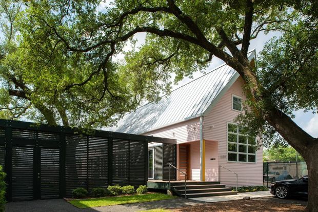 Exterior view of a compact contemporary cottage-style house set beneath mature trees. The pale pink horizontal-clad facade has a steep standing-seam metal gable roof, large white-framed windows, a covered entry, and dark timber or metal screening along the left side. A small paved approach, landscaped planting, and a parked vehicle are visible.
