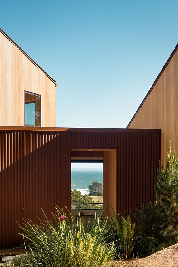 Exterior view of a modern coastal residence composed of timber-clad volumes and a dark vertically corrugated central facade. A framed opening aligns with a visible ocean and coastal landscape beyond, while native planting occupies the foreground. The composition emphasizes warm timber, strong geometric forms, and a sheltered courtyard-like approach under a clear blue sky.
