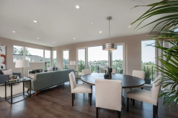 Bright open-plan living and dining area with large corner windows overlooking a leafy suburban landscape. The room features dark timber flooring, pale taupe walls, a grey sofa, a round dark dining table with upholstered chairs, a fireplace, recessed ceiling lights, and a suspended metallic pendant above the dining area. Tall indoor greenery frames the right side.