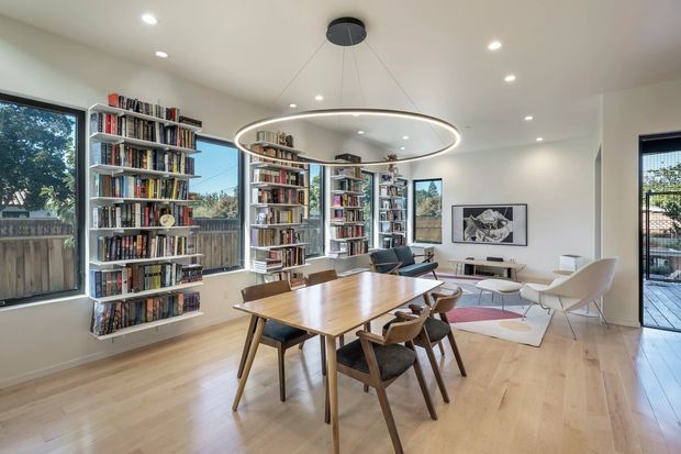 Bright, open-plan library and dining area with extensive built-in book shelving, a timber dining table and chairs, pale timber flooring, contemporary lounge seating, and large windows opening toward an outdoor deck. A sculptural circular pendant light and recessed ceiling lights provide illumination, while neutral finishes create an airy, refined atmosphere.