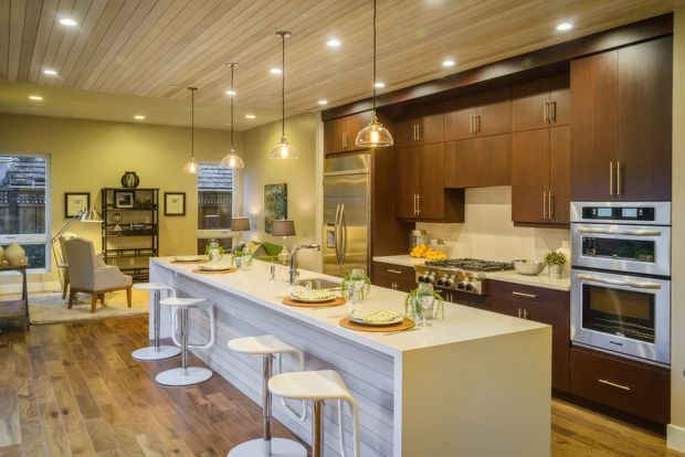 Warm, spacious open-plan kitchen with a large white island and breakfast seating, dark timber cabinetry, integrated stainless-steel appliances, pendant lights, and a timber-lined ceiling. A dining/living area is visible beyond the kitchen.