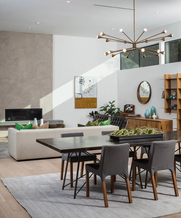 A bright, open-plan living and dining area with a contemporary mid-century-inspired interior. The foreground features a dark rectangular dining table with upholstered charcoal dining chairs and a low planter centerpiece. Behind it is a light modular sofa, a black fireplace or TV inset into a textured grey feature wall, a timber sideboard, open timber shelving, decorative ceramics, and an oval wall mirror. A sculptural brass-and-glass starburst chandelier hangs beneath a high white ceiling, while large upper windows bring in strong natural light.