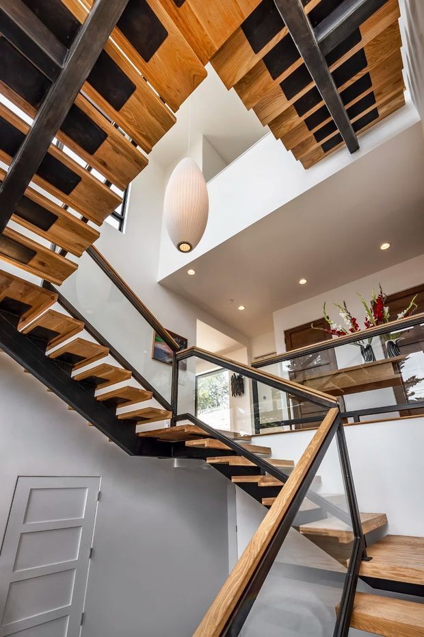 Interior view of a contemporary stairway rising through a tall open void. The staircase has floating timber treads supported by dark metal stringers, with matching timber handrails and black-framed glass balustrades. A sculptural pendant light hangs centrally beneath exposed timber ceiling elements, while white walls, recessed downlights, and upper-level glazing create a bright, airy vertical space.
