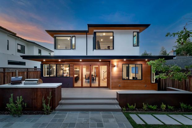 Twilight exterior view of a contemporary two-storey residence with a flat roof and clean white upper-level walls over a warm timber-clad lower level. Large glazed doors open onto a raised deck and outdoor kitchen area, with dark rendered boundary walls, paved landscaping, and timber fencing.