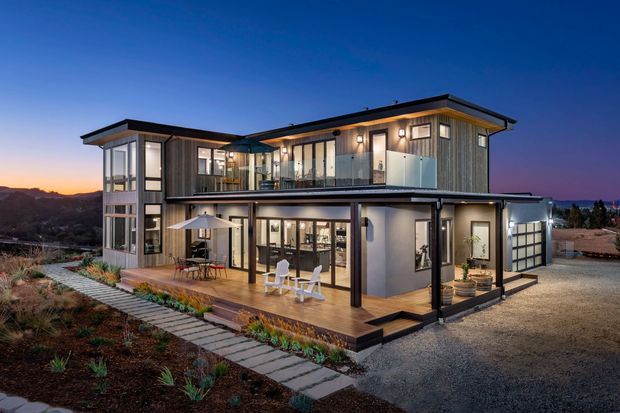 Twilight exterior view of a contemporary two-storey home with a compact flat-roofed form, extensive floor-to-ceiling glazing, dark metal framing, vertical timber-look cladding, and a covered ground-floor terrace. The house sits on a landscaped, sloping site with paved paths and gravel surrounding it.
